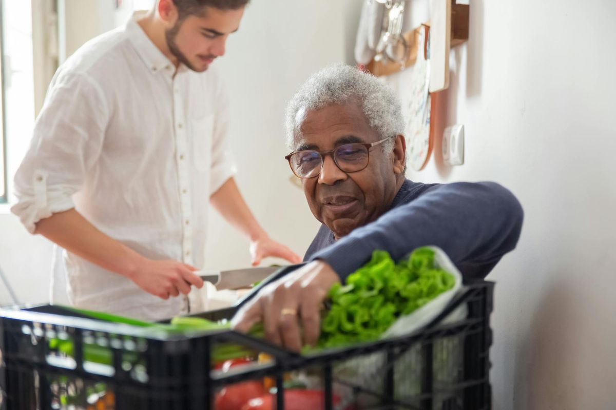 A companion helps an older man unpack fresh groceries at home